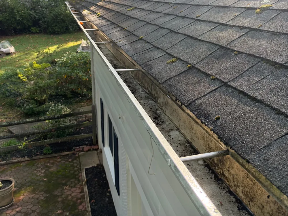Open gutter with dirt and debris built up inside along a shingle roof edge, before guard installation